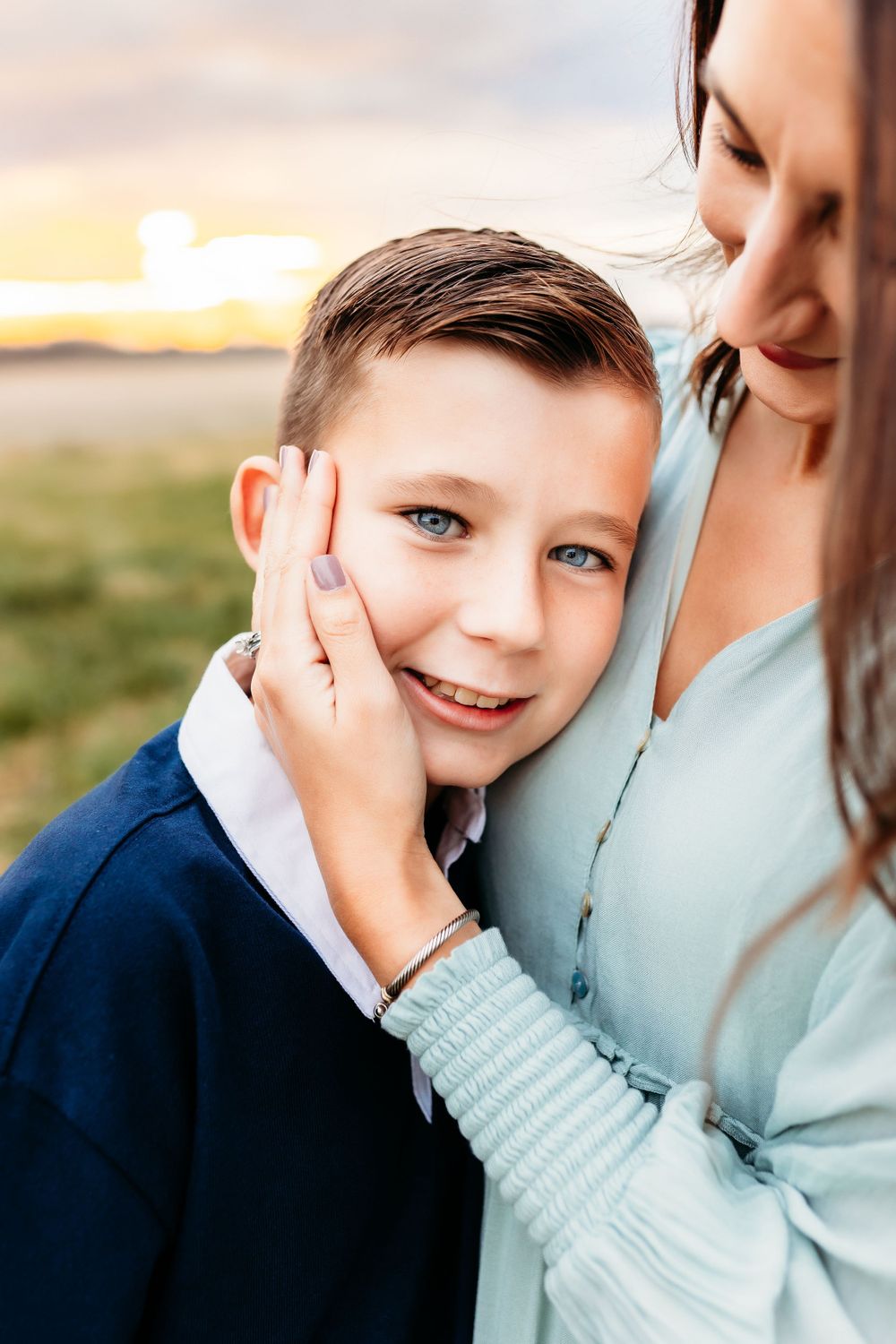 A smiling blue-eyed boy leaning into his mothers embrace during a golden sunset portrait.