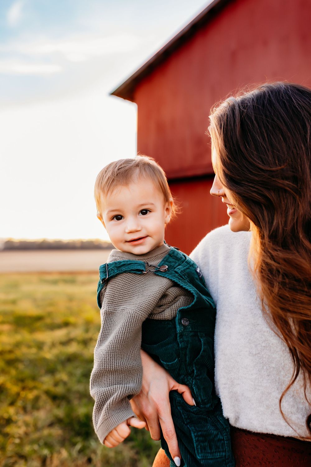 A smiling baby boy in green overalls held by his mother near a red barn at golden hour.