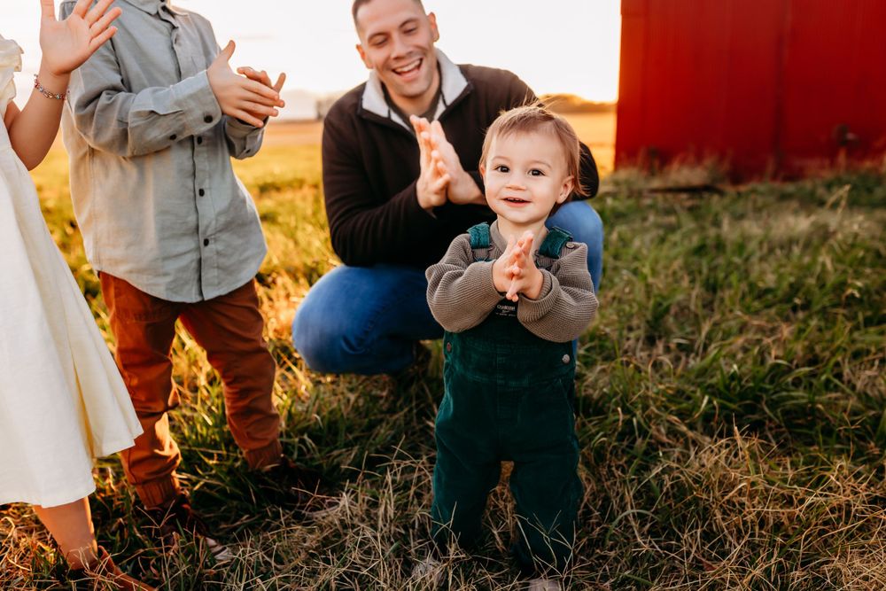 A happy toddler boy clapping hands with his father crouching beside him near a red barn.