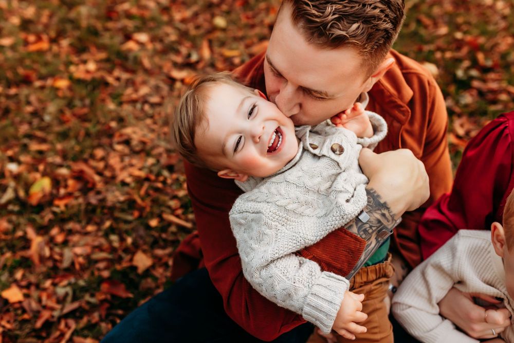 A laughing toddler girl being hugged and kissed by her father outdoors in fall foliage.