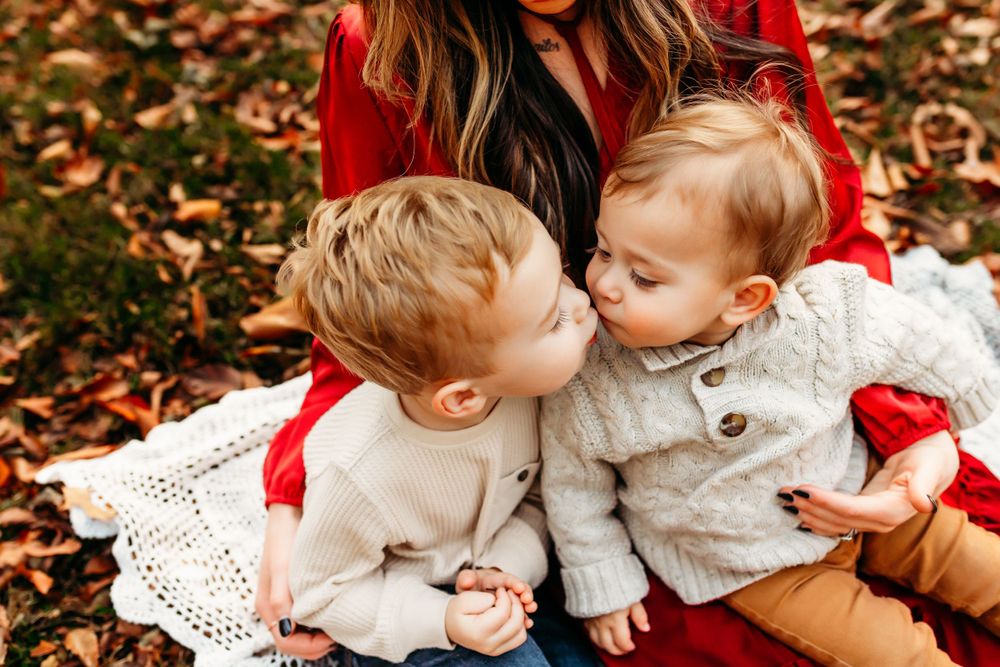Two young siblings sharing a sweet kiss while sitting on a blanket surrounded by autumn leaves.