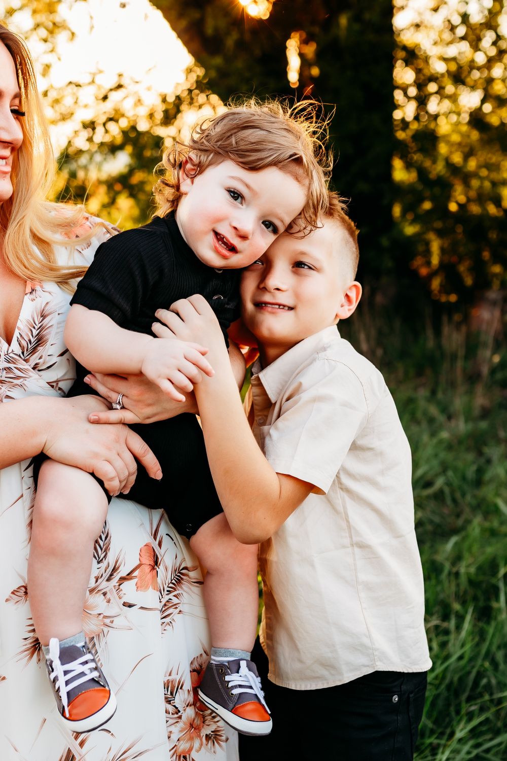 A mother holding two young boys close together in a warm golden outdoor family portrait.