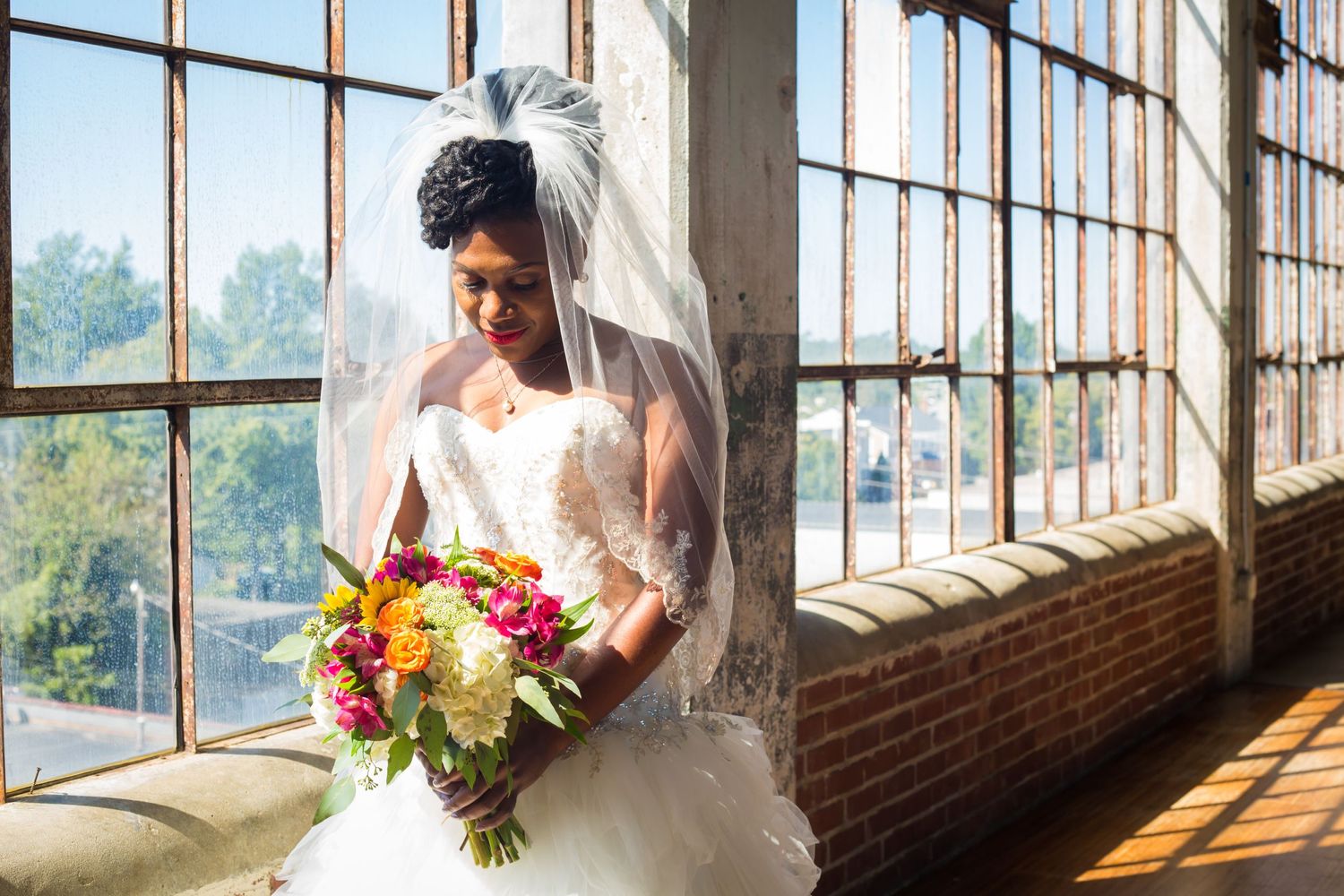 A bride in a white gown holds a vibrant autumn bouquet while standing by sunlit industrial windows.