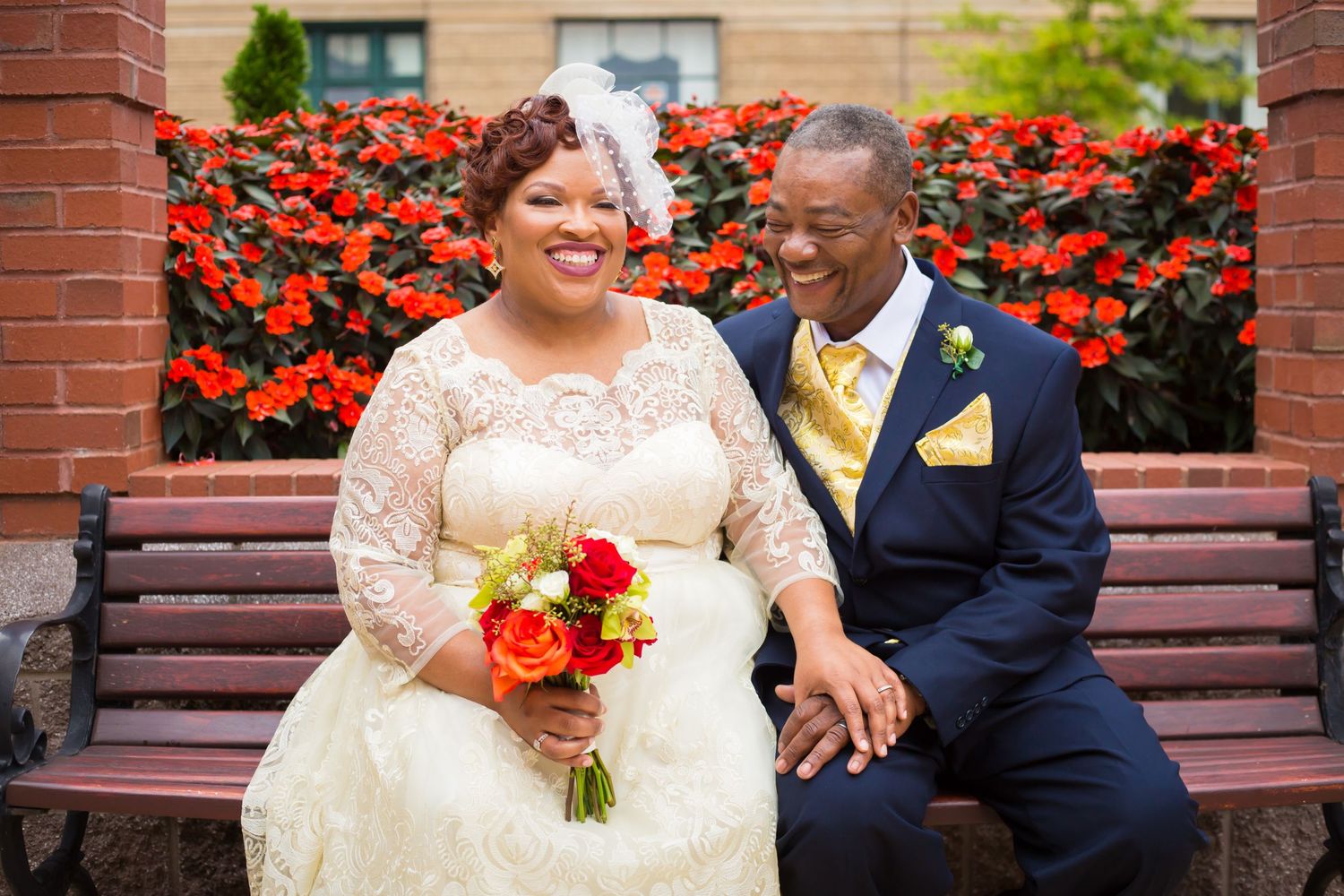 Couple shares a happy moment on bench with vibrant red flowers in background.