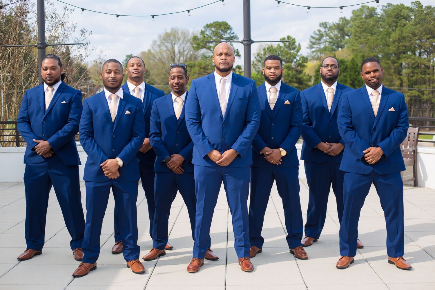 Groomsmen stand in coordinated royal blue suits with brown shoes outdoors.