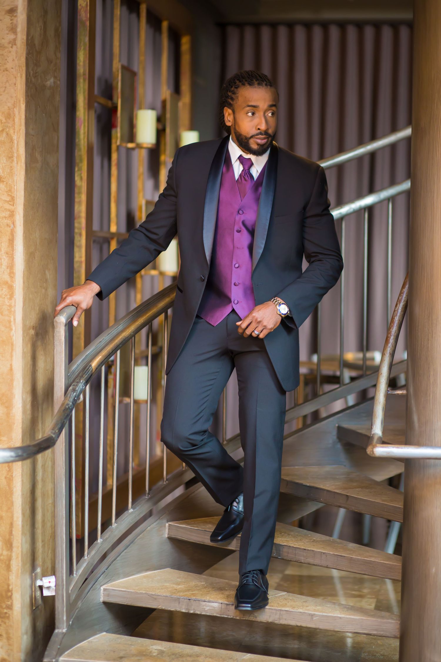 A groom descends a spiral staircase wearing a dark suit with purple vest and tie.