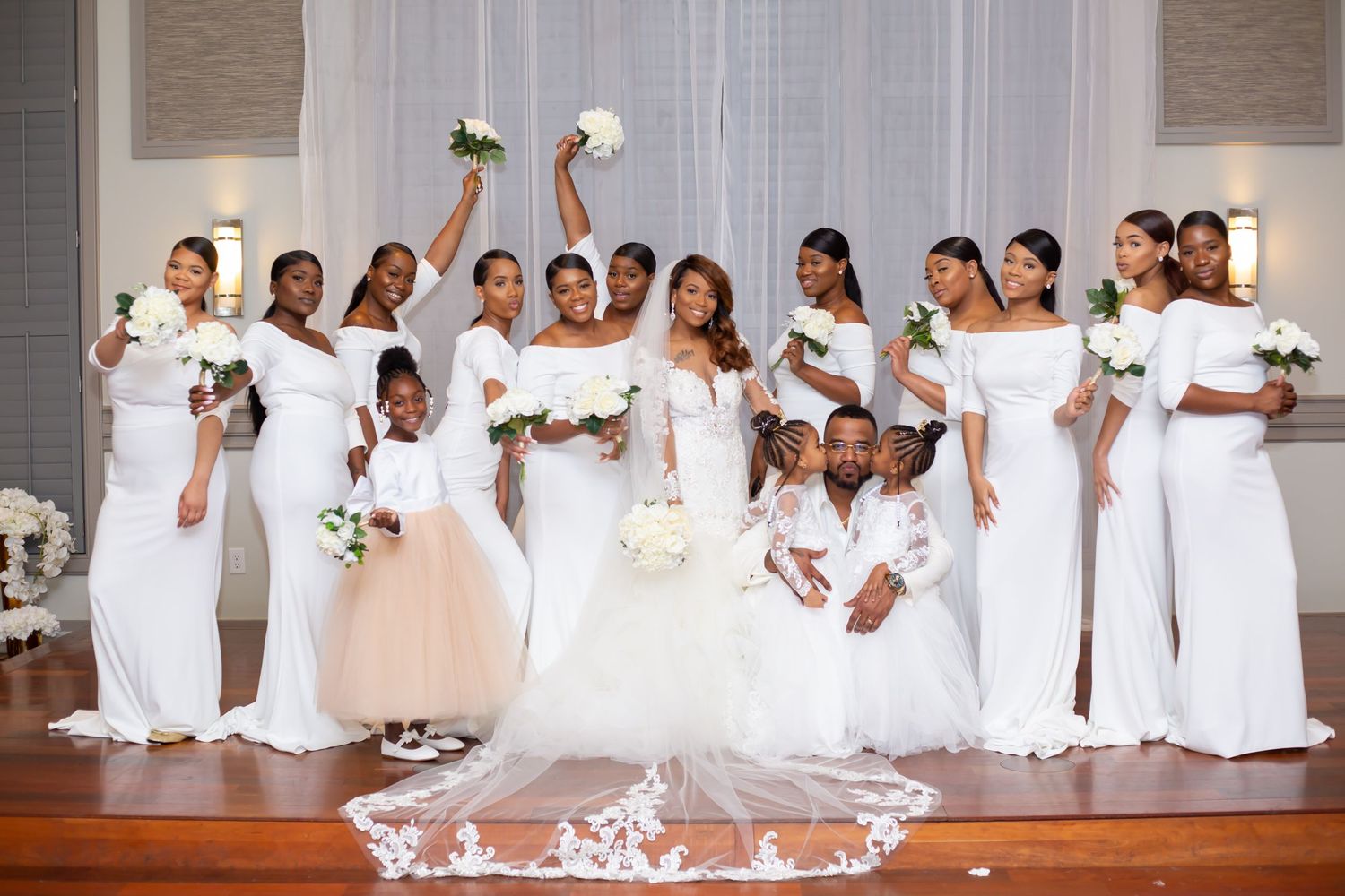 Large wedding party in white dresses poses together for celebratory group photo.