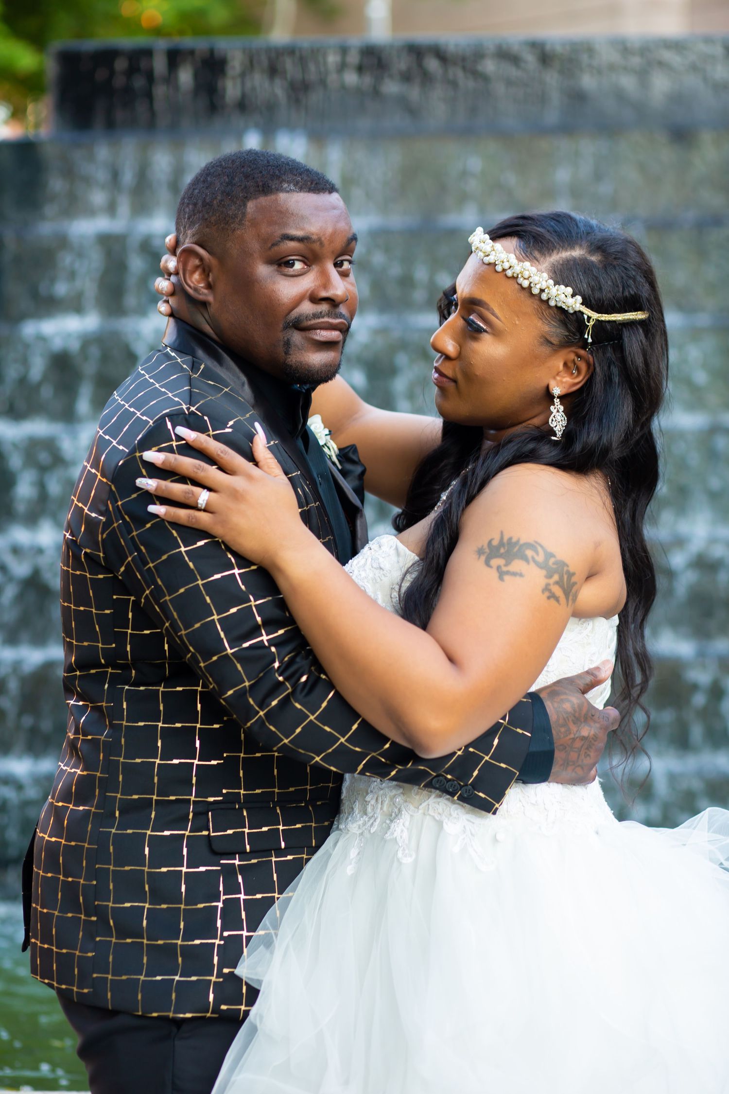 Couple embraces in formal wear against water wall backdrop outdoors.