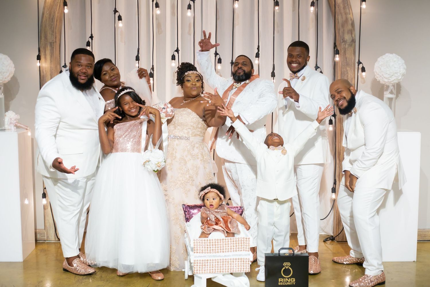 Wedding party poses in all white attire against a neutral backdrop with hanging lights and balloons.