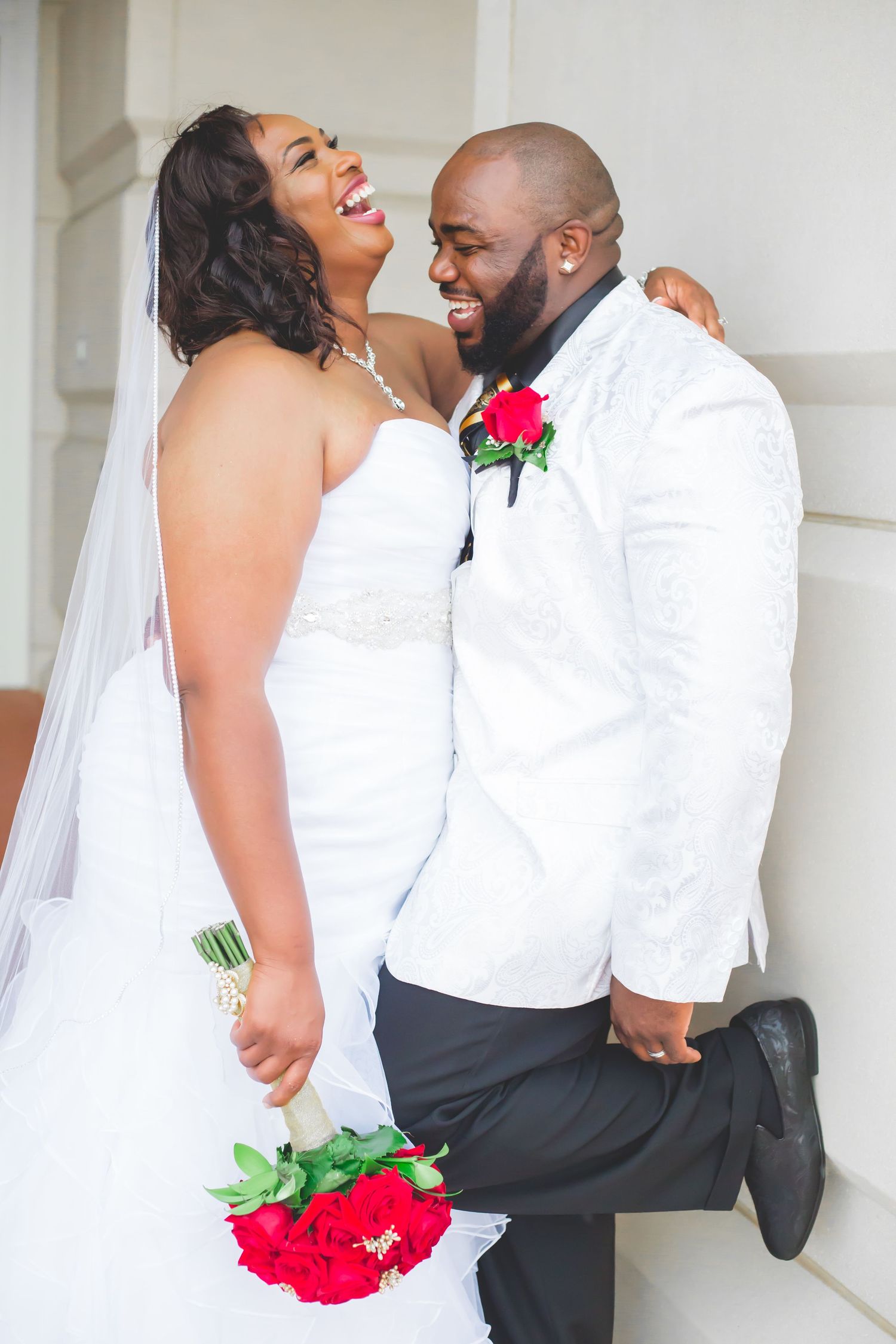 Newlyweds share a joyful moment in white attire with red floral accents.
