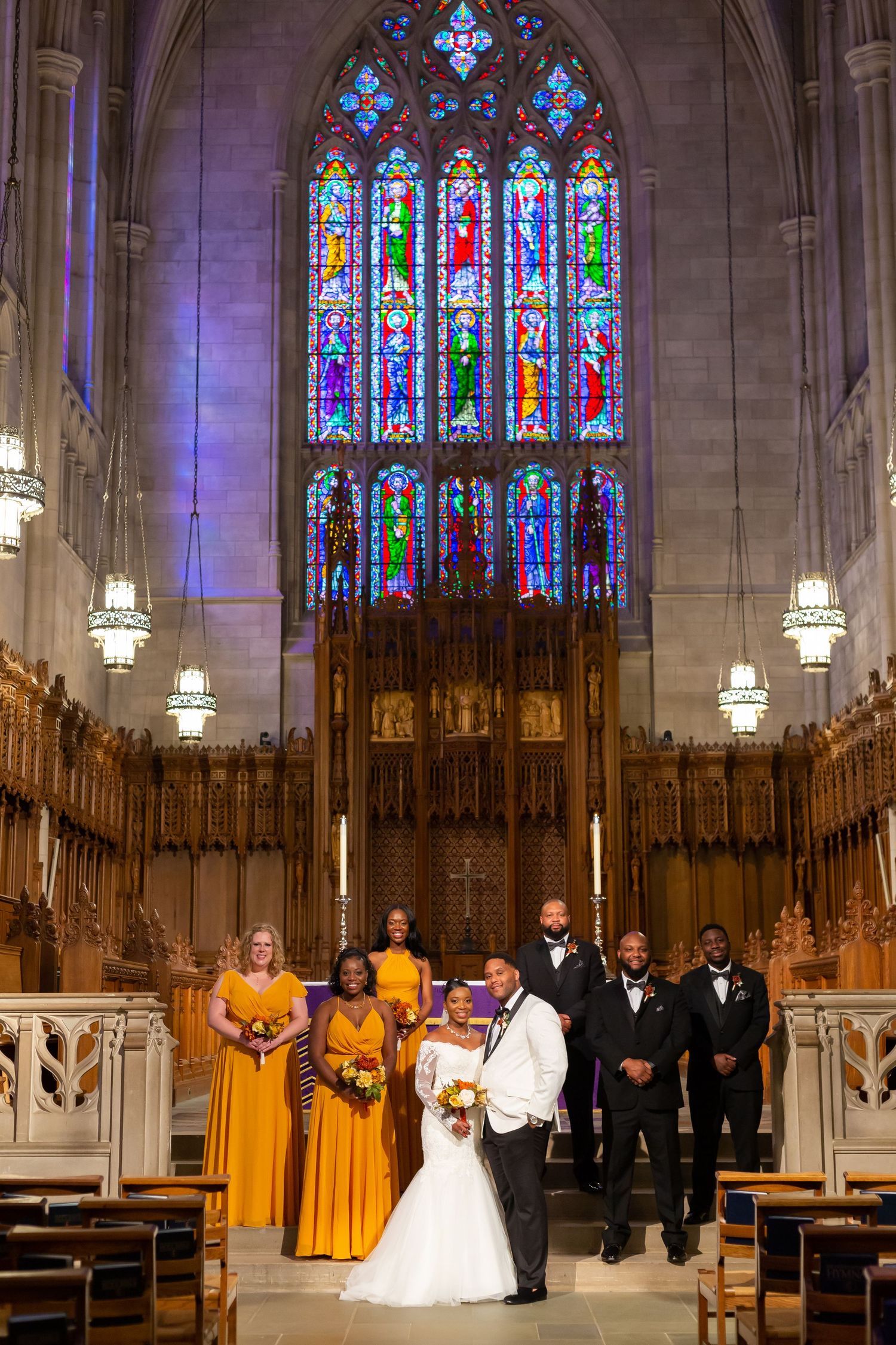 Wedding party poses in gold and white attire under stunning stained glass windows in a cathedral.