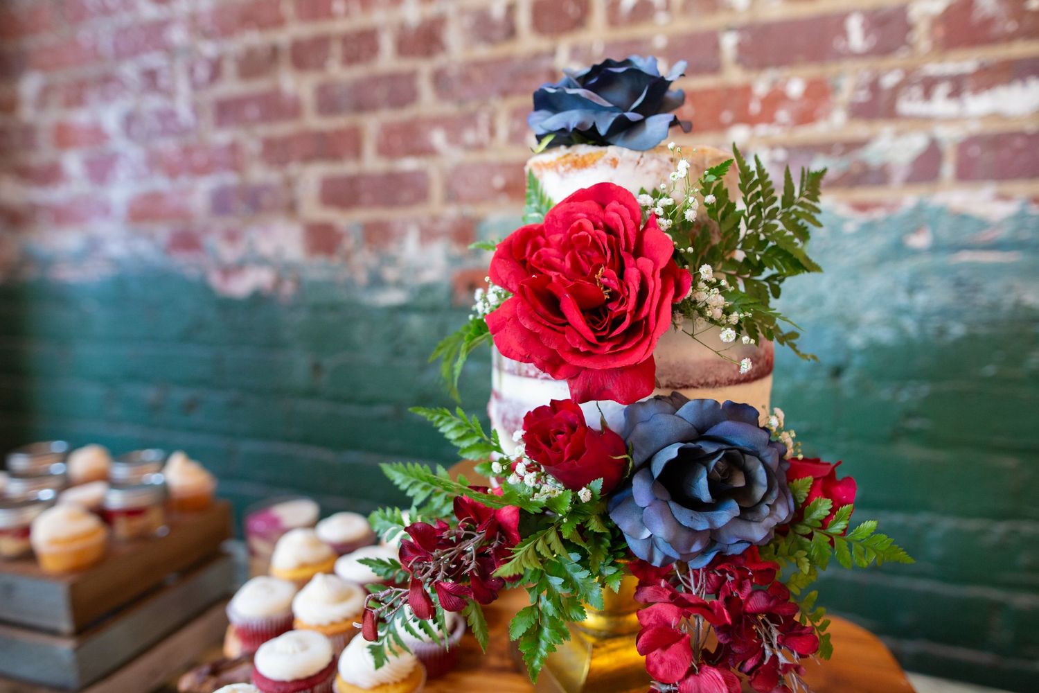 Wedding cake decorated with deep red roses and blue succulents against rustic brick backdrop.