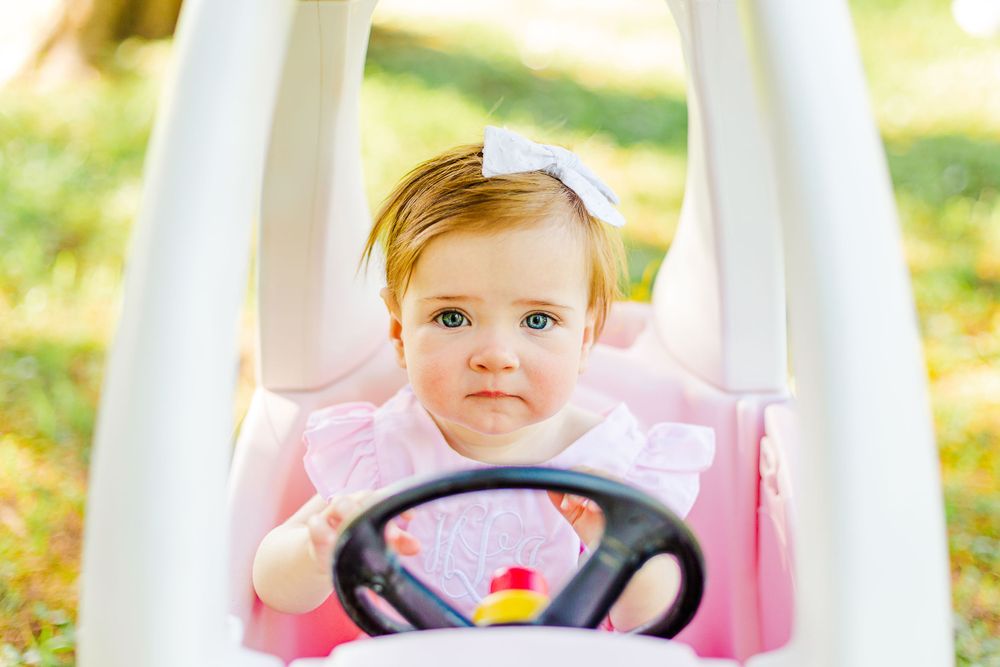 Baby wearing pink outfit grips steering wheel of white toy car in sunny garden.