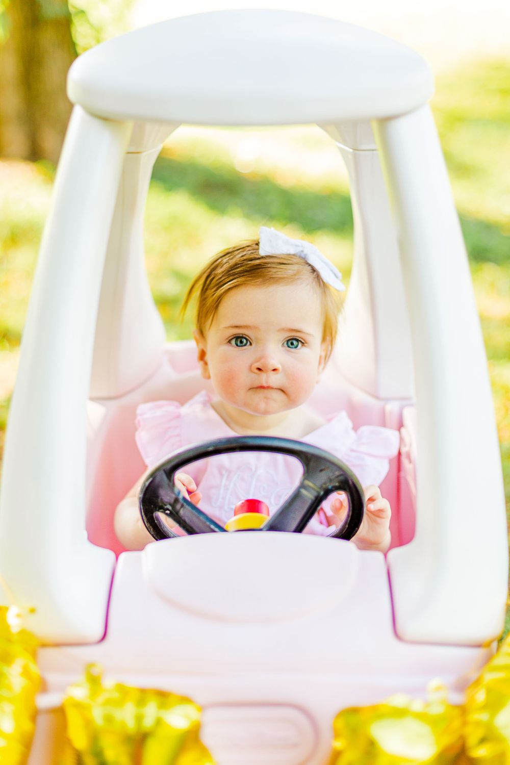 Baby in pink outfit holds steering wheel of white toy coupe car in sunlit garden.
