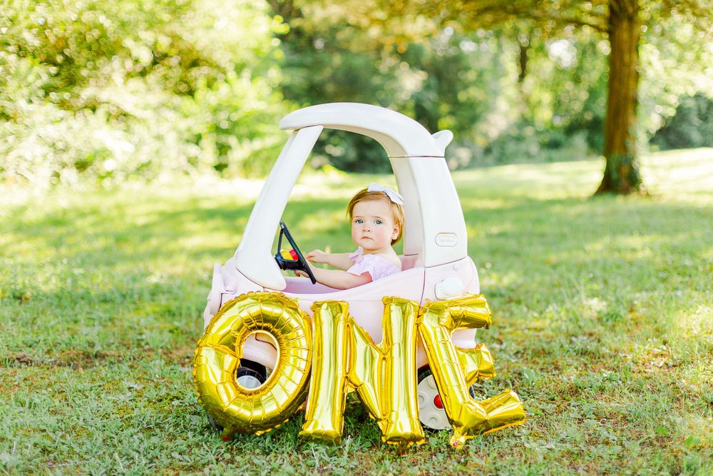 A first birthday photo featuring a small child in a white toy car with gold letter balloons spelling 'ONE'.