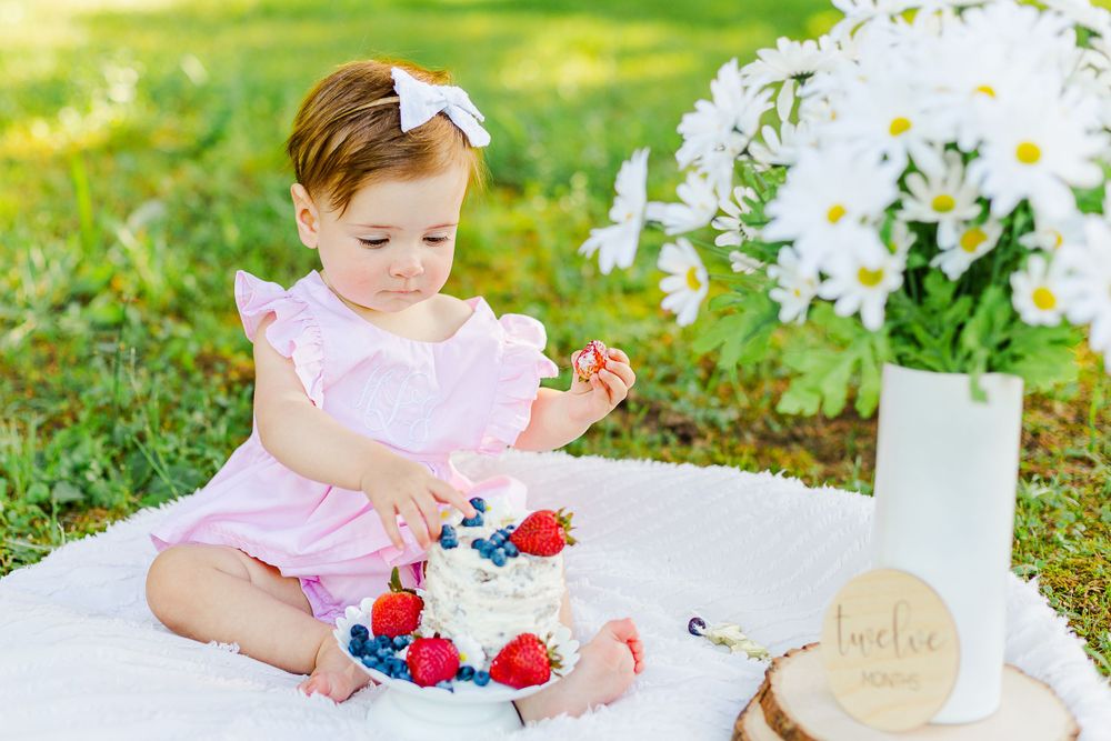 Pink-dressed baby seated on white blanket with berry-topped cake and daisies.