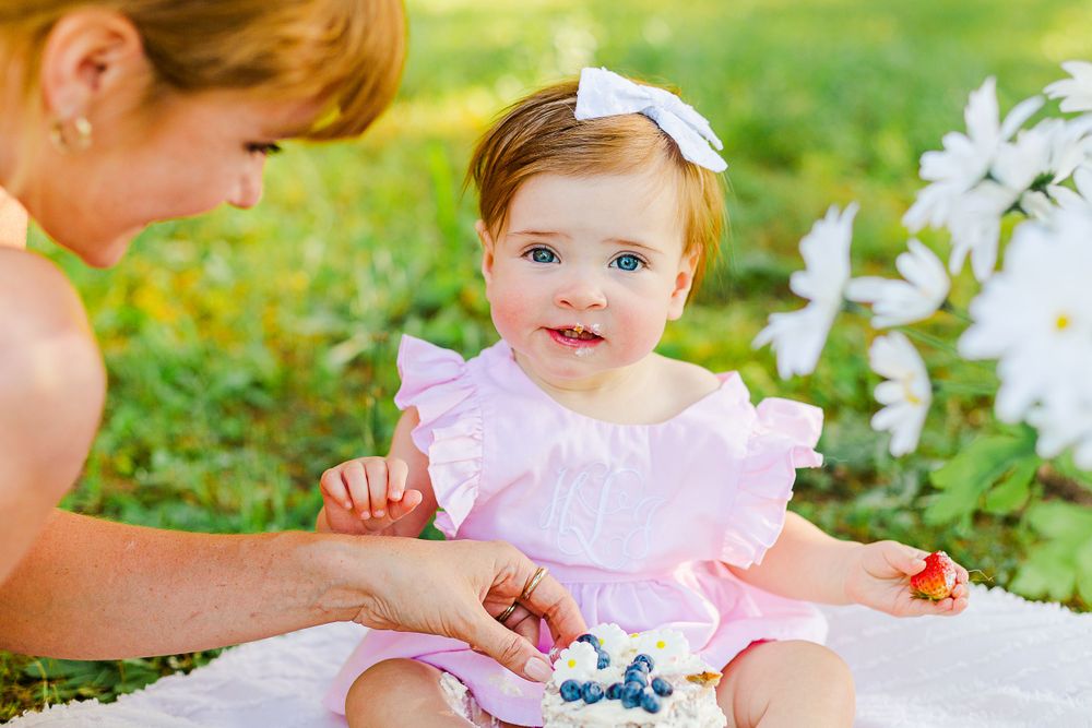 A baby in a pink ruffle dress sits on a white blanket exploring snacks and treats in an outdoor garden setting.