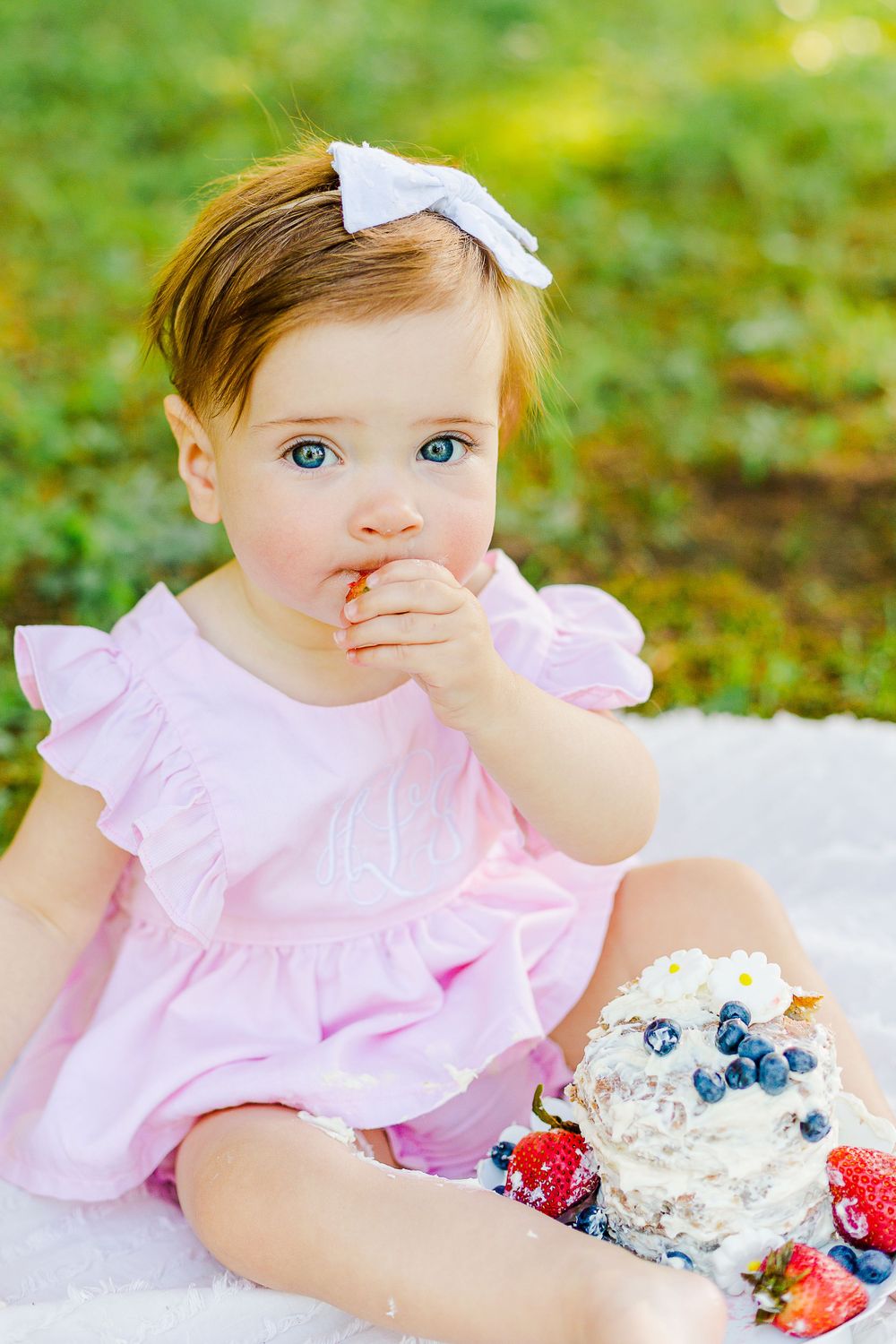 A curious little one in pink dress tastes dessert while sitting on white blanket with decorated cake outdoors.