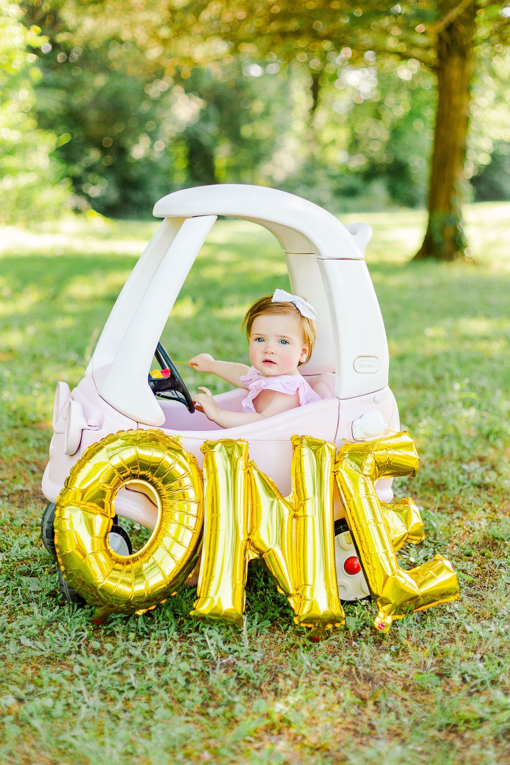 A toddler sits in a white toy car with gold 'ONE' balloons for first birthday photos.