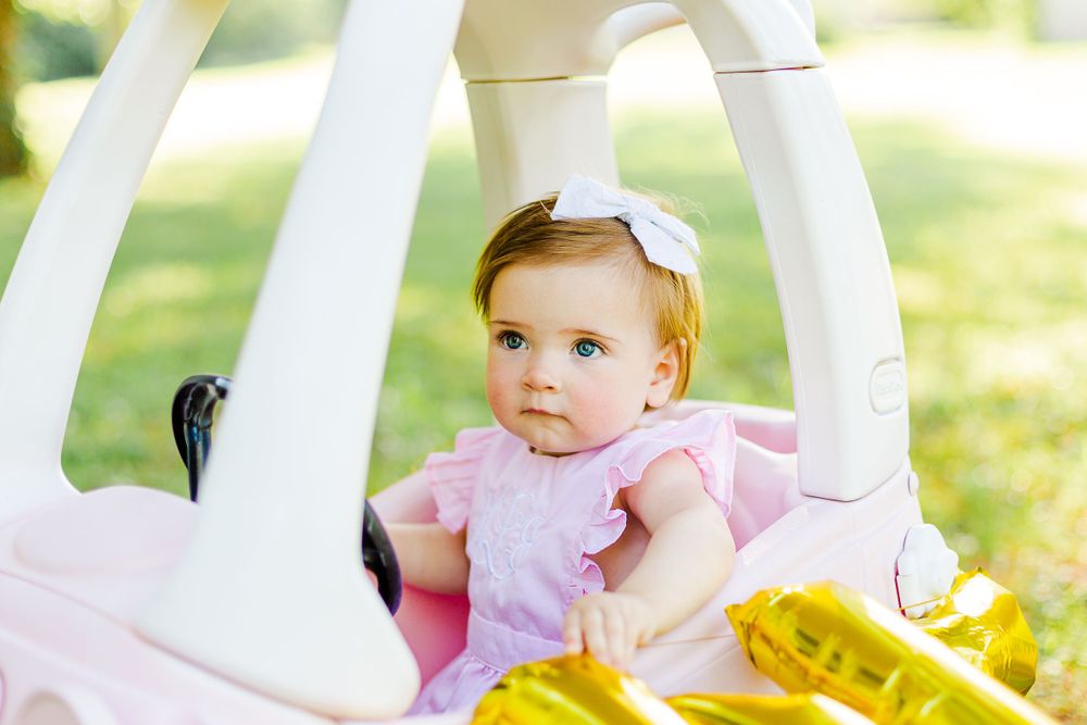 Pink-dressed baby sits in white toy cozy coupe car with gold balloon decorations on sunny grass.