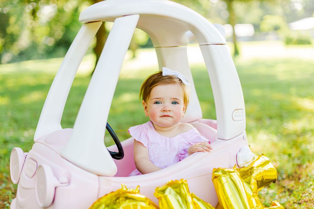 Baby wearing pink dress sits in white toy car with gold balloons in outdoor garden setting.