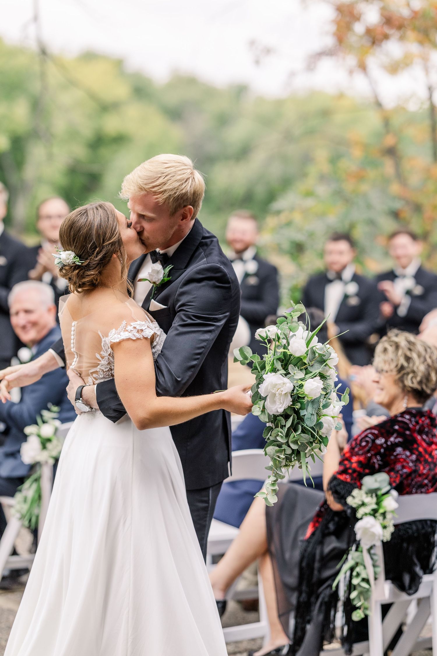 Couple embraces during outdoor wedding ceremony with floral arrangements and guests.