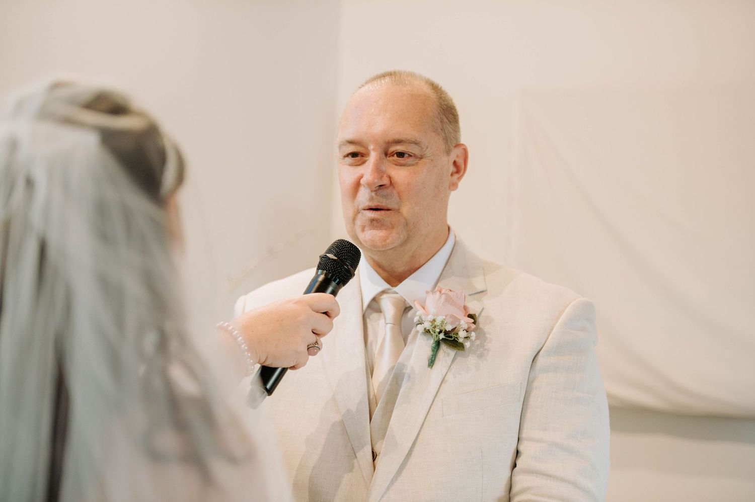 A person in a light beige suit speaks into a microphone during a wedding ceremony.