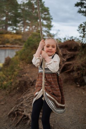 Child in patterned dress holding rope swing on forest path.
