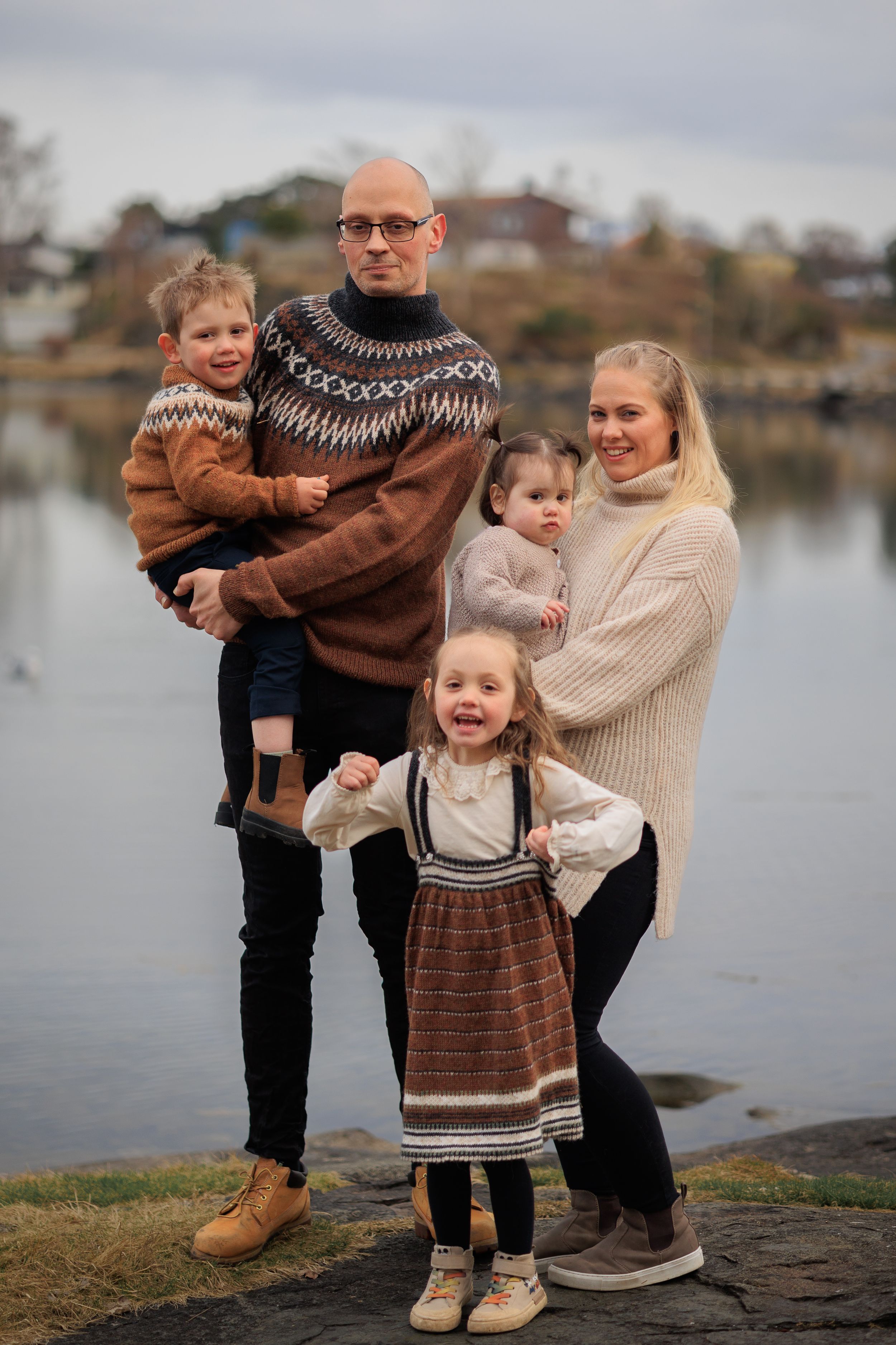 Family wearing coordinated knit sweaters posing together by lake on cloudy day.