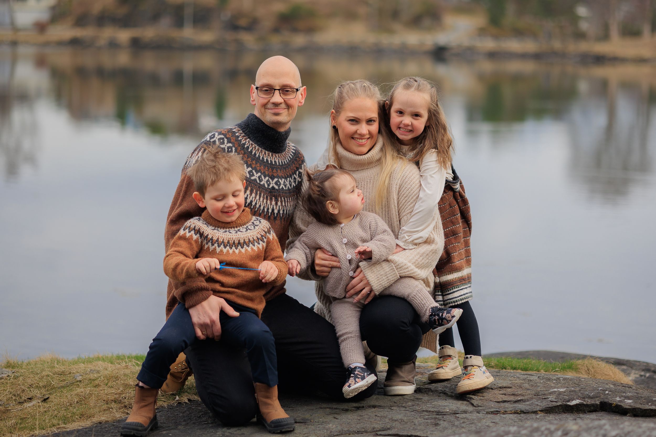 Family wearing coordinated winter sweaters sitting together on rocky lakeside.