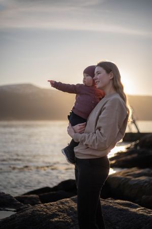 A parent and child share a tender moment on rocky shoreline during sunset.