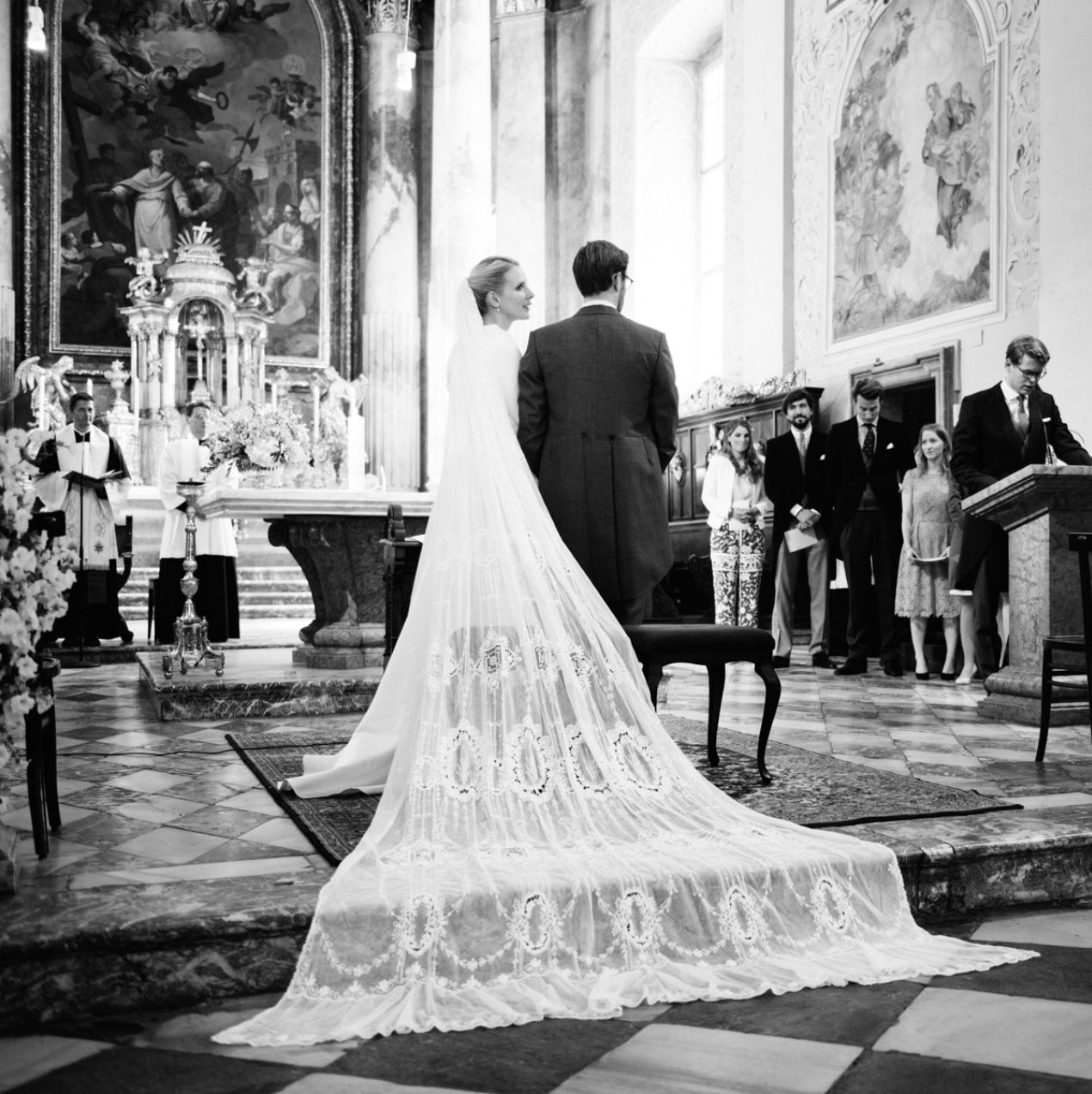 Black and white photo of a wedding ceremony in an ornate church with a long lace train.