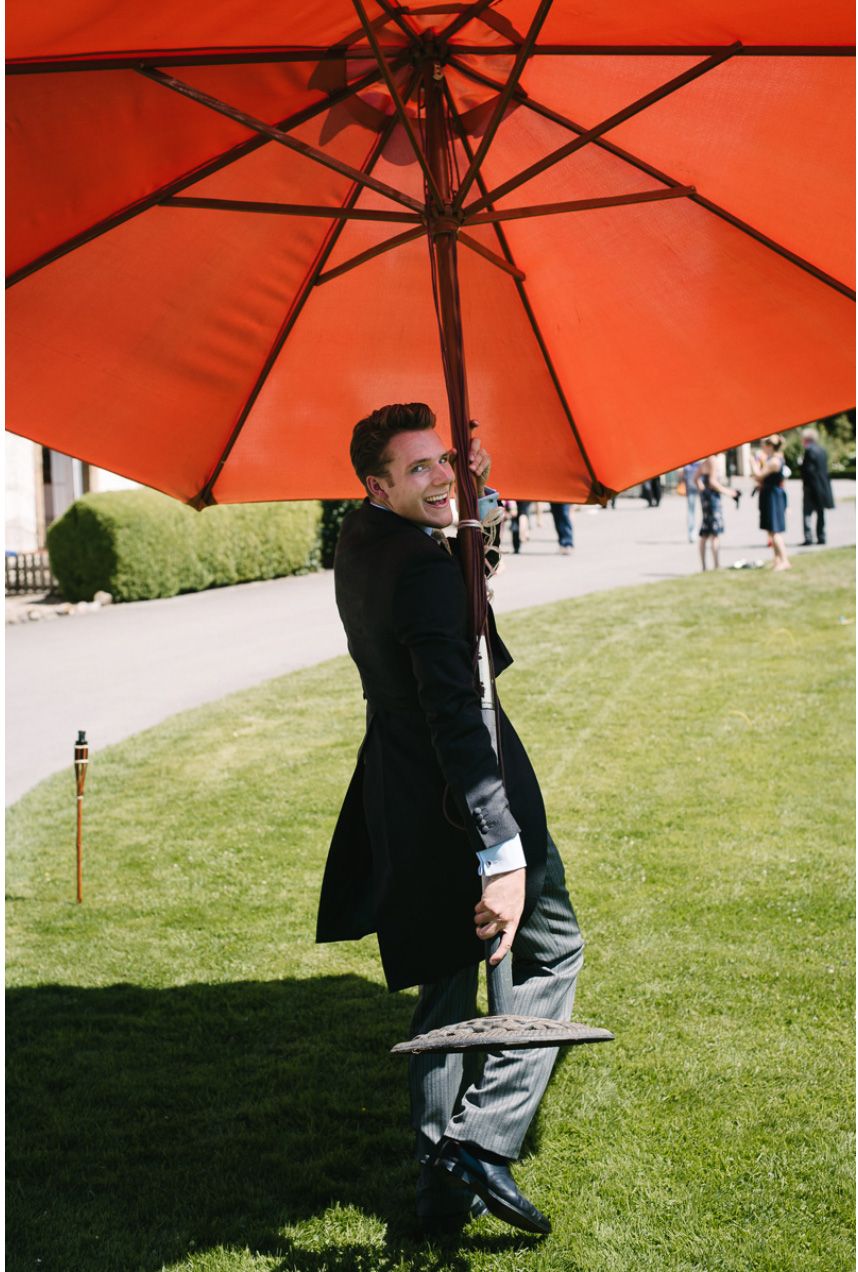 Person in formal attire playfully holds a large red umbrella on a sunny lawn.