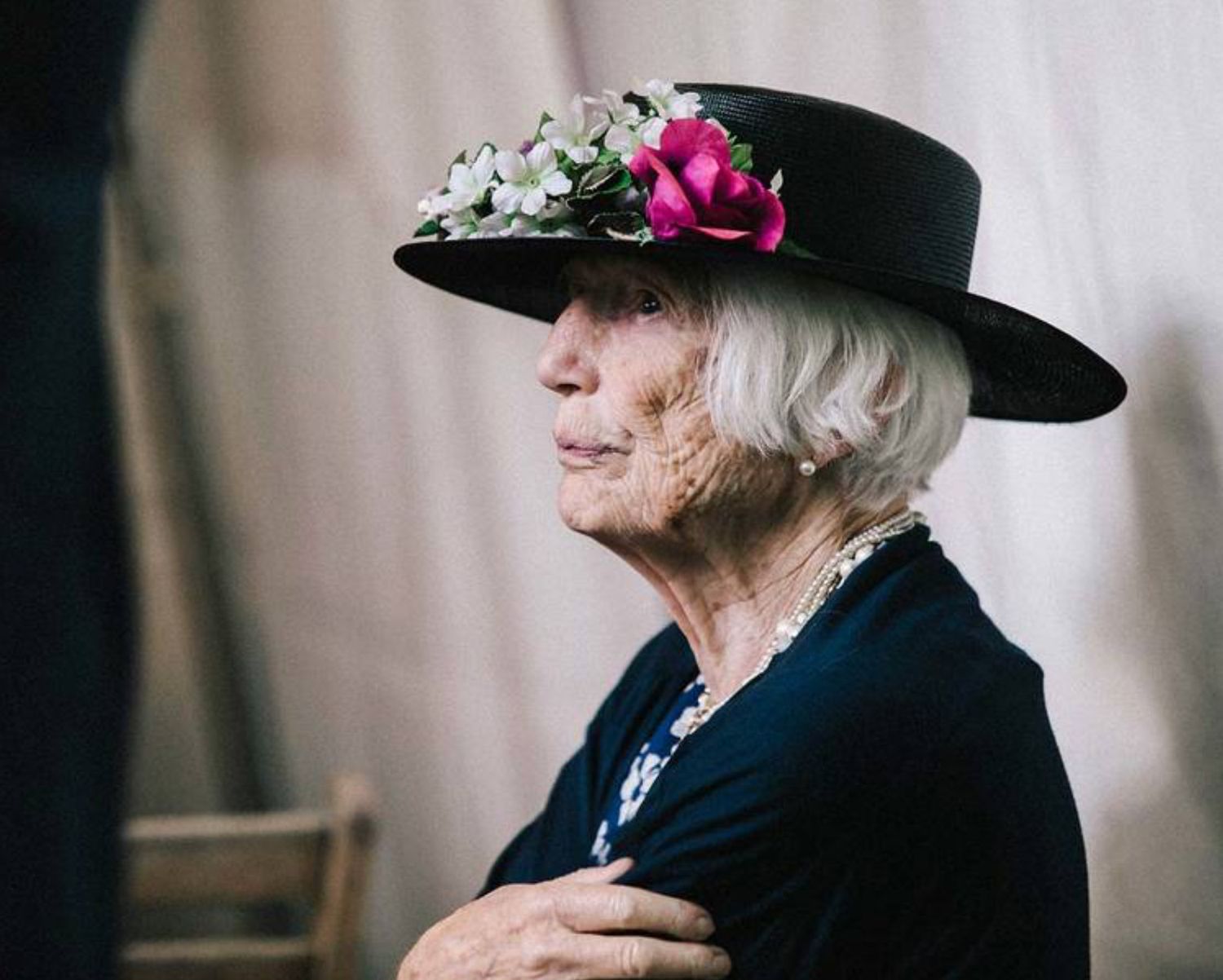 A woman wearing a decorated black hat with pink flowers gazes to the side.