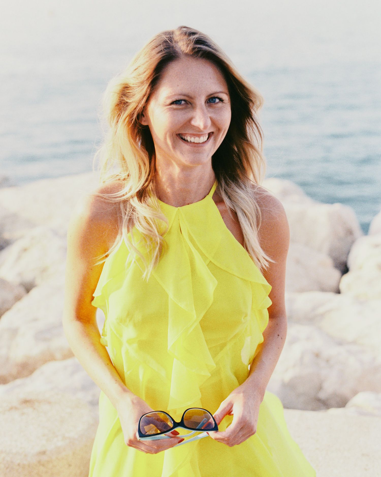 Woman in bright yellow summer dress poses on rocky beach near water.