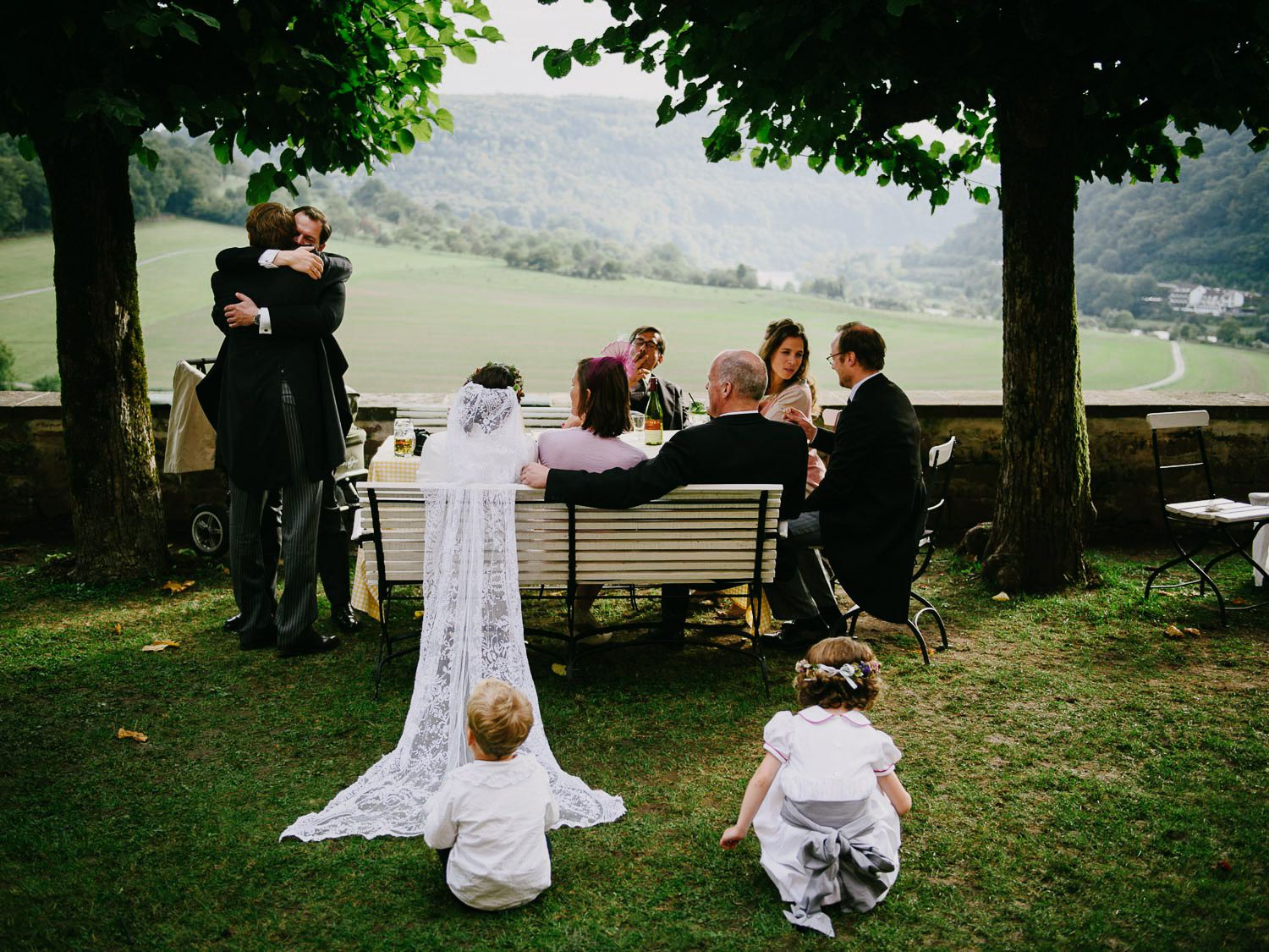 Wedding scene with small children watching as guests gather under trees on a hillside.