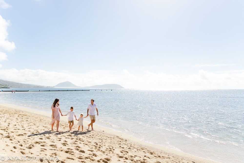 A family walks along a scenic beach shoreline with mountains visible in the distance.