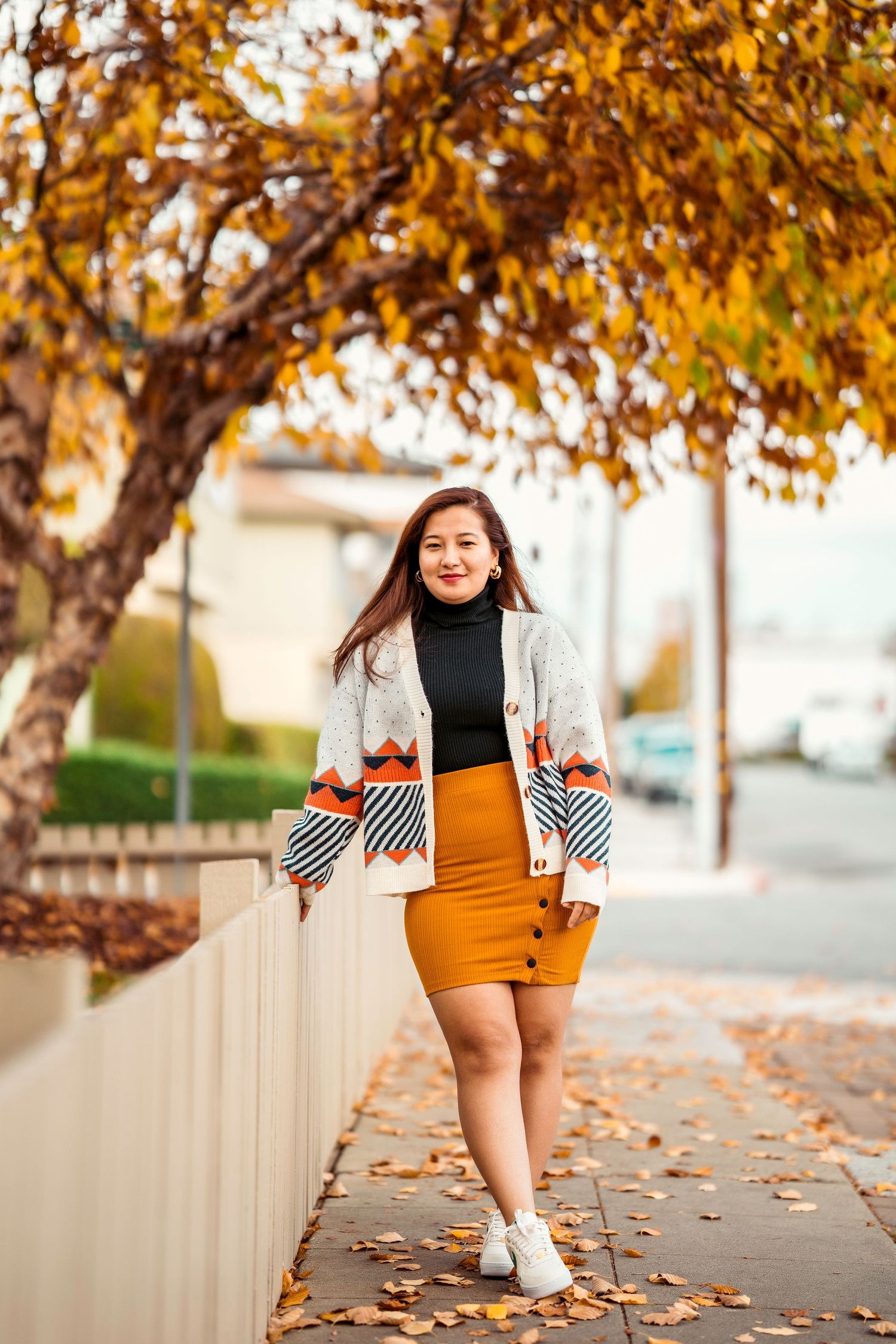 Woman in autumn attire with patterned cardigan and orange skirt poses against fall foliage background.