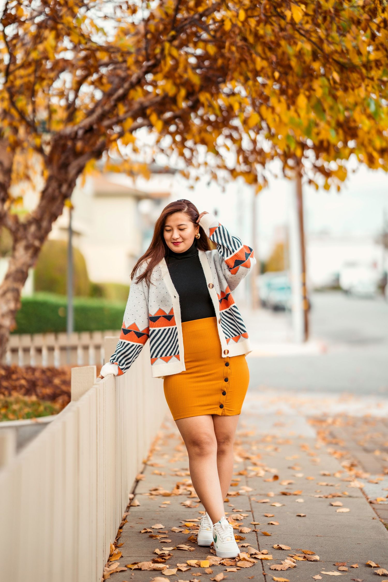 Stylish fall fashion outfit featuring geometric pattern cardigan and mustard pencil skirt on urban sidewalk with autumn leaves.
