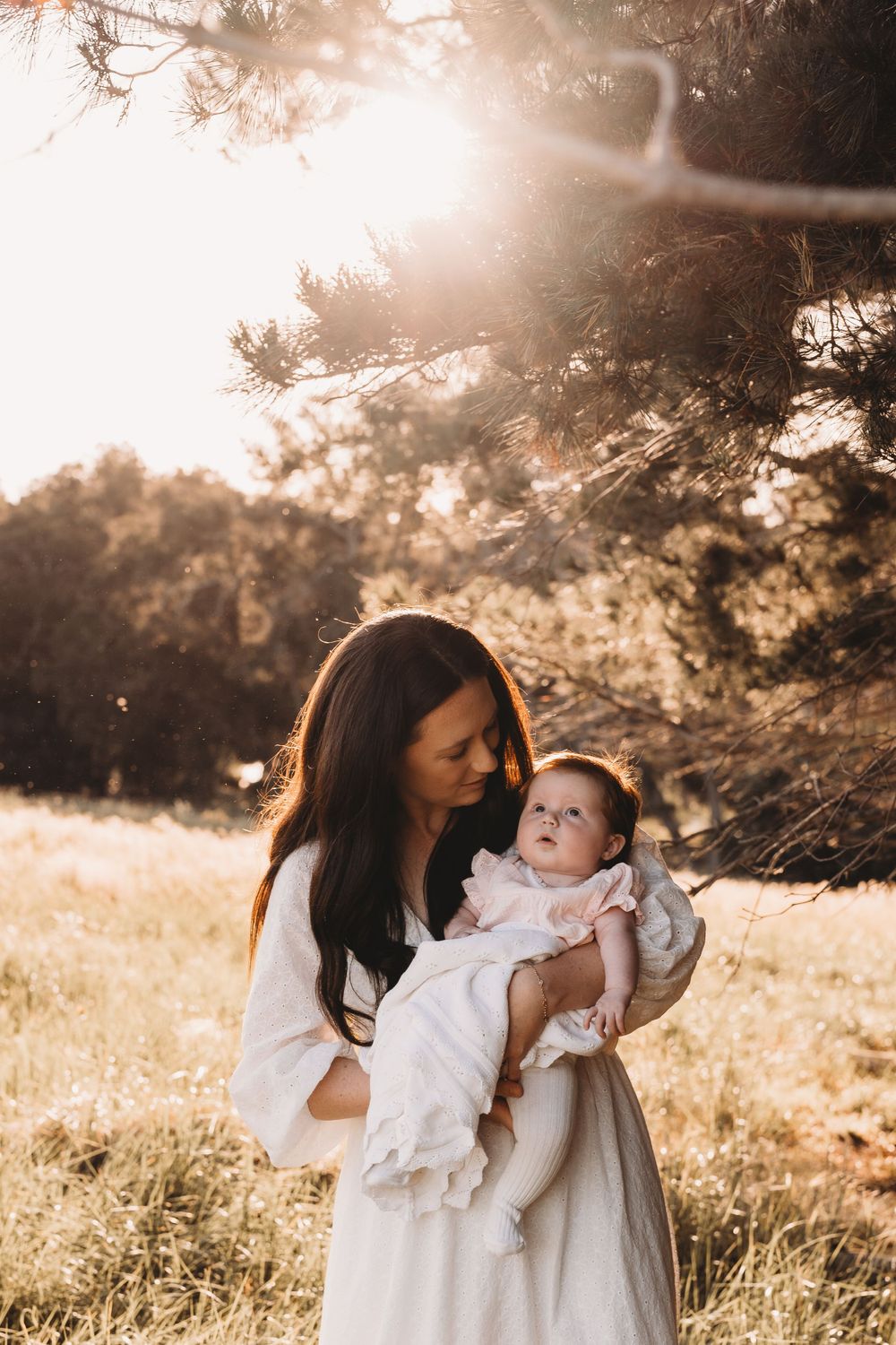 Dark-haired mother holding her infant daughter in soft golden sunlight outdoors.
