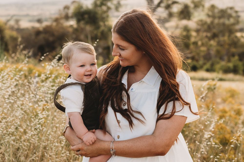 Brunette mother holding her happy baby boy in a sunlit field of wildflowers.