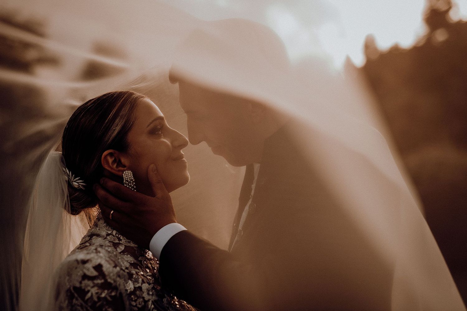 Groom tenderly cupping brides face as golden veil wraps around them in soft warm light.