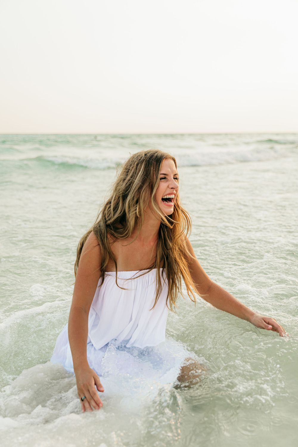 Person in white dress laughing while standing in ocean waves.