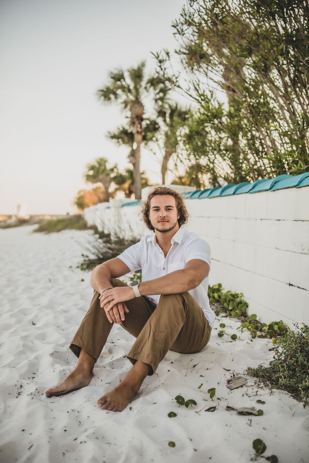 Person sitting barefoot on white wall near palm trees at beach.