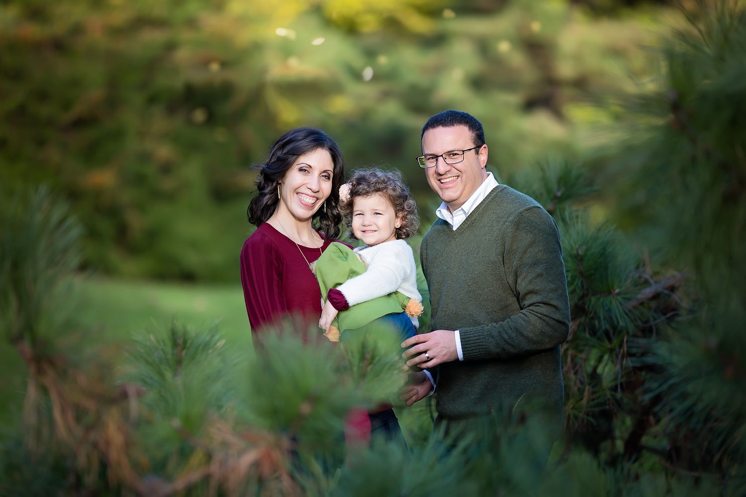 Happy family of three posing among lush green pine trees in a nature portrait.