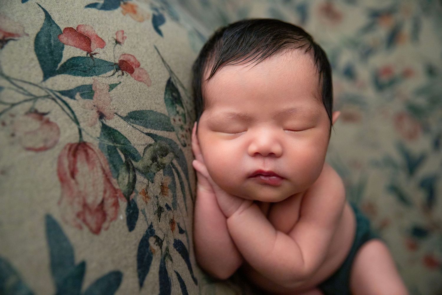 Newborn baby sleeping peacefully on a floral backdrop with hands tucked under chin.