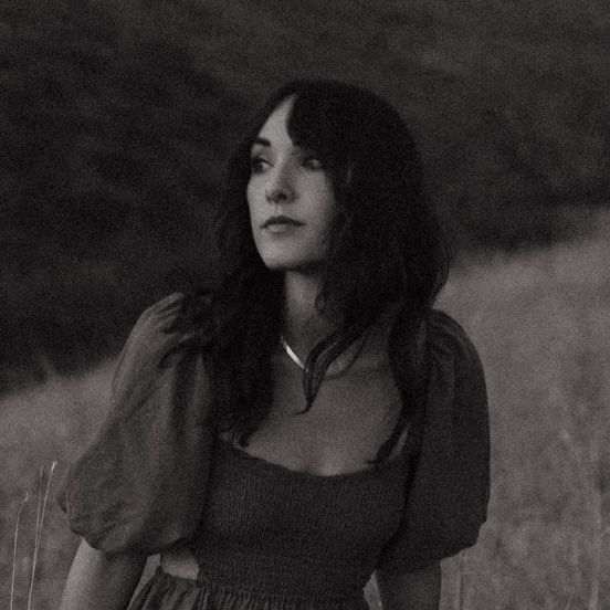 Black and white portrait of a young woman with dark hair standing in an open field.