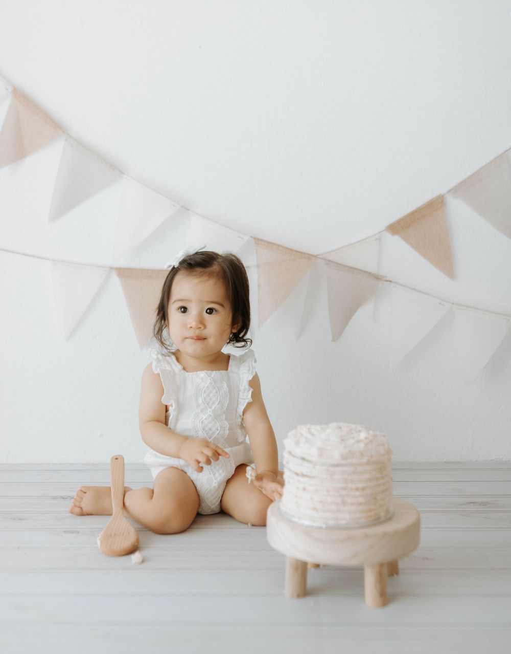 Toddler in white dress sitting next to small white cake with bunting decorations.