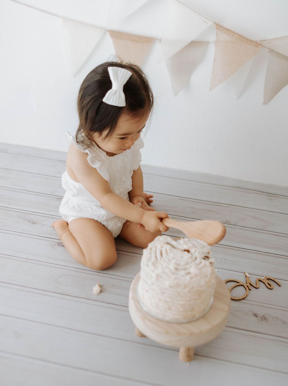 A small child in white lace romper examines a white cake on wooden stand under party banners.