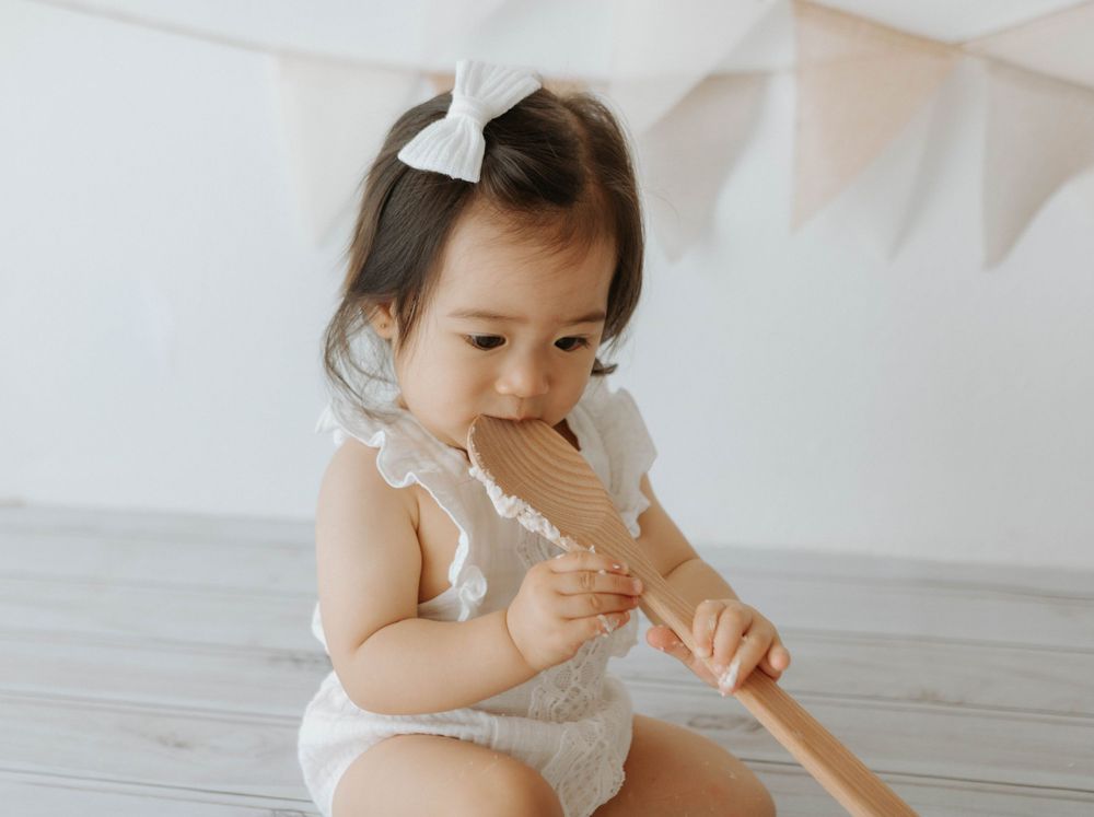A toddler in white outfit plays with a wooden spoon during a cake photoshoot.